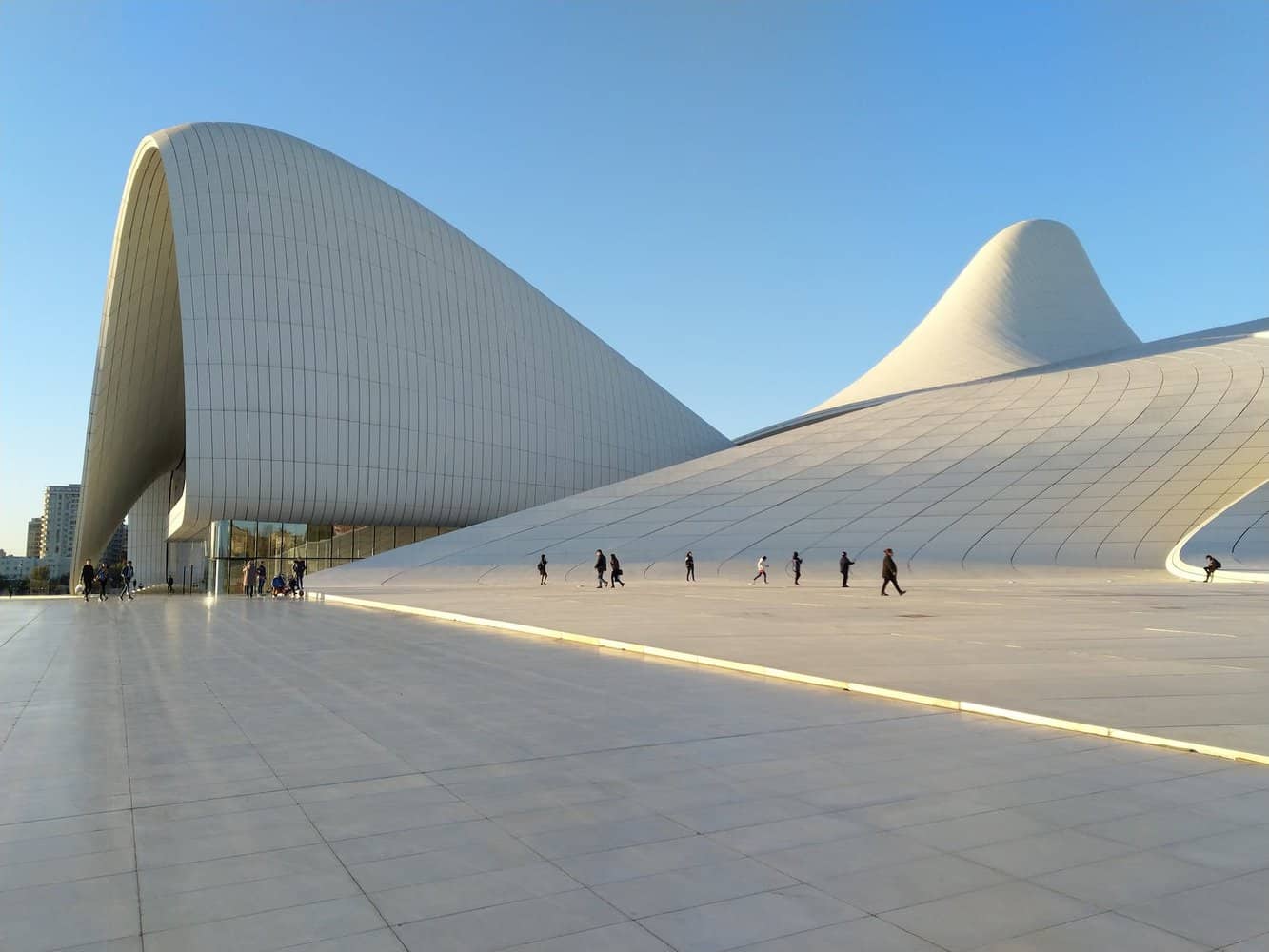 photo of people walking near building designed by zarhar hadid, a found architecture. Giant white slopes slip and slide forming a unique shape of buidling. Making BAKU oc