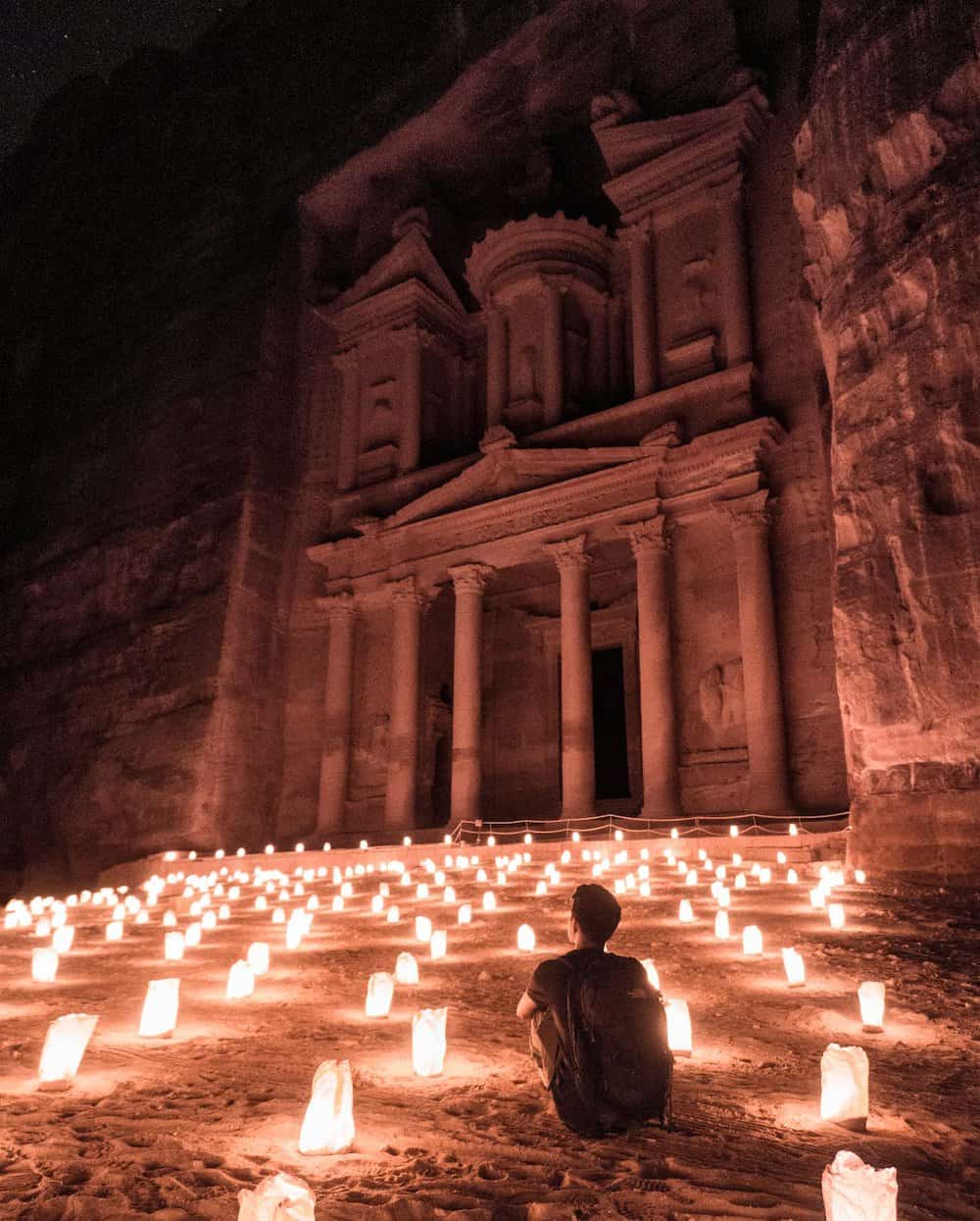 a man sitting outside of the famous petra building carved into red stone sliffs. a famous one of the biblical sites