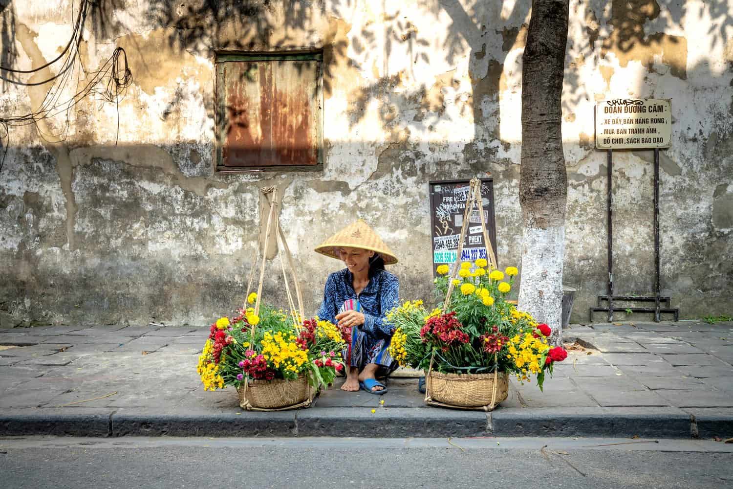 delighted young asian female seller sitting on street near baskets of fresh flowers