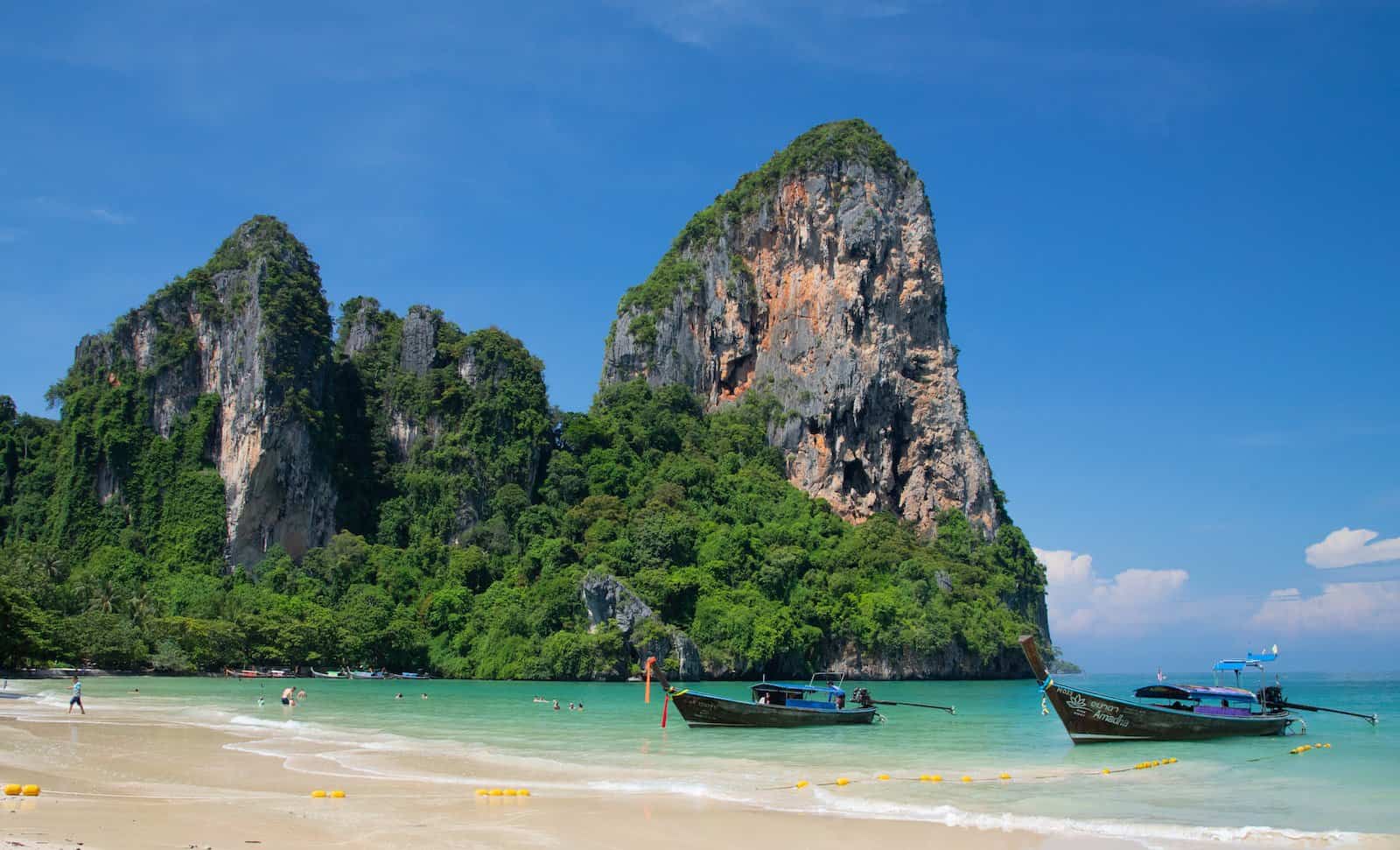 a beach with boats and a large rock formation in the background with Railay Beach in the background. bali vs thailand 